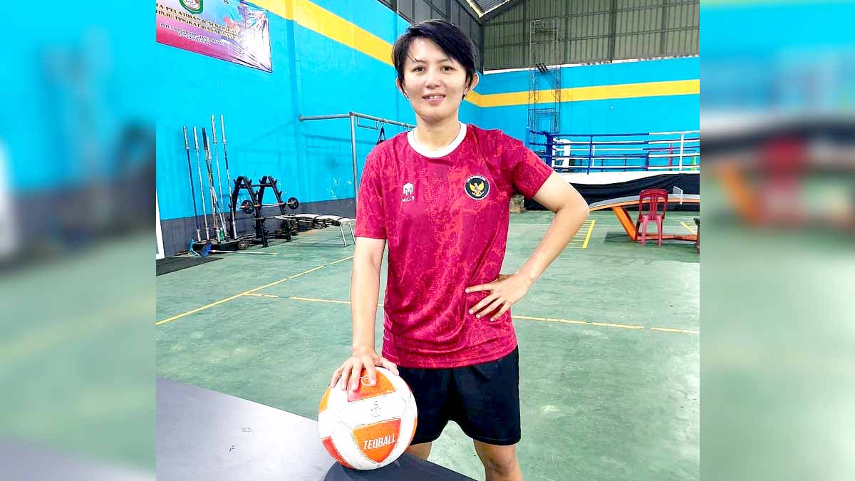 Brigpol Akyko Kapito, Seorang atlet perempuan tampak berdiri di dalam area latihan indoor, mengenakan jersey merah bergambar lambang burung Garuda. Ia memegang sebuah bola Teqball di salah satu sisi meja latihan, dengan latar peralatan fitness dan ring tinju yang berada di belakangnya. Foto: IST
