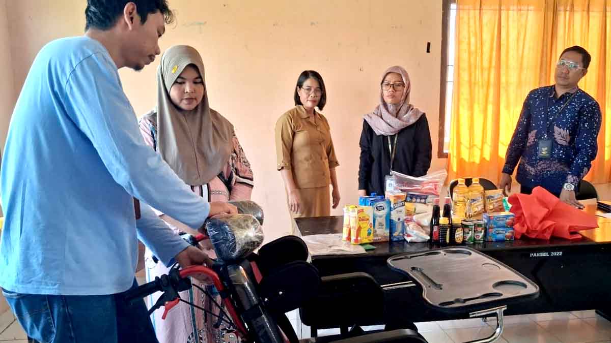 Keterangan Foto: Seorang petugas terlihat menjelaskan penggunaan alat bantu berupa kursi roda kepada seorang penerima manfaat, sementara tiga perempuan dan seorang pria lain menyaksikan proses penyerahan bantuan. Di atas meja tampak berbagai paket bantuan kebutuhan dasar dan perlengkapan dukungan mobilitas. Foto: Diskominfo